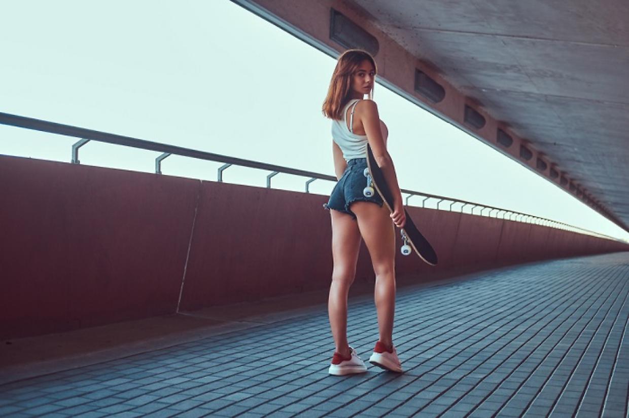Back view of a sensual young skater girl dressed in shorts and t-shirt holds a skateboard and looking at the camera.